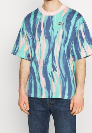Mężczyzna w za dużym T-shircie Adidas z abstrakcyjnym wzorem pociągnięć pędzla w kolorach niebieskim, zielono-niebieskim i różowym, w połączeniu z ciemnoniebieskimi dżinsami.