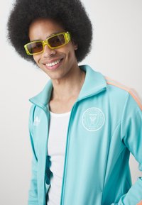 Akvamarin trackjakke med orange striber, lynlåskrave og hvidt logo-patch. Båret over en hvid tanktop, parret med gule solbriller.