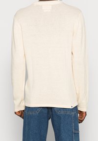 Maglione a maniche lunghe di colore crema realizzato in morbido tessuto a maglia. Presenta un colletto a costine e una cucitura discreta sul retro. L'orlo è leggermente curvato.