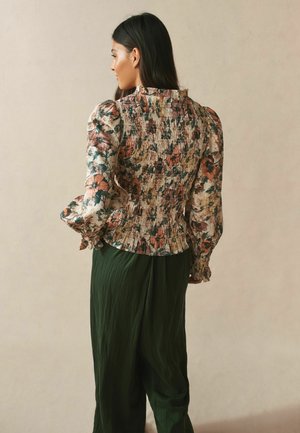 Woman with long dark hair wearing a floral puff-sleeve blouse and dark green wide-leg pants, standing turned slightly away against a plain background.