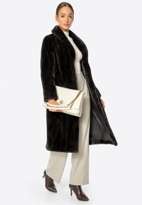 Donkerbruine nepbontjas met een sjaalkraag, lange mouwen en een zachte textuur. Gecombineerd met beige wijde broekspijpen en een witte clutch met gouden hardware.