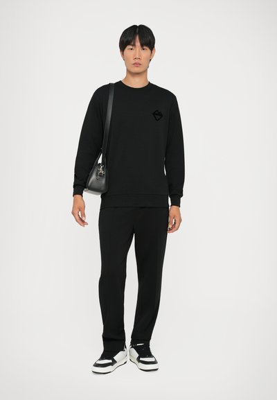 Sweatshirt nero con un logo discreto, pantaloni neri abbinati e sneakers con dettagli bianchi. Tessuto morbido con una vestibilità rilassata. Borsa a tracolla inclusa.