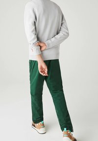 Osoba w jasnoszarym swetrze, ciemnozielonych spodniach i beżowych trampkach, stojąca z rękami złożonymi za plecami na białym tle.