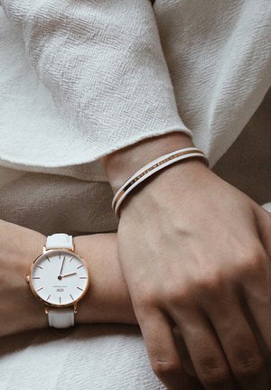 Bela usnjena ura z rožnatimi zlatimi detajli na zapestju. Na drugem zapestju je viden tanek bel in zlat bangle uhani. Teksturirana ozadje.
