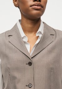 Άτομο που φοράει καφέ και λευκό καρό σακάκι πάνω από λευκό πουκάμισο, εμφανίζεται από τον λαιμό έως το μέσο του κορμού σε απλό φόντο.