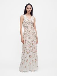 Femme aux longs cheveux foncés portant une robe blanche longue sans manches avec une broderie florale rose, debout sur un fond blanc uni.