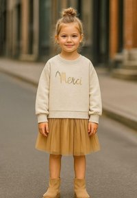 Beiger Sweatshirt mit "Merci" in Gold, kombiniert mit einem hellbraunen Tüllrock und hellbraunen Stiefeletten. Die Stoffe sind weich und strukturiert.
