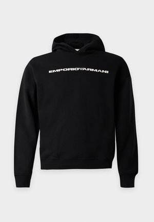 Mees, kes kannab musta kapuutsiga särki valge "EMPORIO ARMANI" tekstiga, musti dressipükse, valgeid tossusid ja tumedaid päikeseprille, seisab valgel taustal.