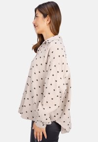 Light beige button-up shirt with black heart pattern, textured fabric, relaxed fit, and dropped shoulders, shown from the side.