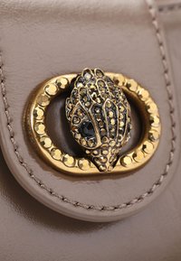 Accessorio in ecopelle beige con chiusura dorata, decorato con dettagli neri e dorati, bordato da piccoli chiodini dorati.