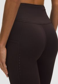 Leggings cu talie înaltă din țesătură maro închis, având un buzunar pe lateral și un model subtil cu puncte de-a lungul cusăturii. Textură netedă și croială elastică.