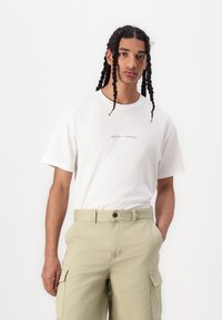 White cotton t-shirt with "WELCOME TO PARADISE" text, paired with beige cargo pants featuring side pockets and a relaxed fit.
