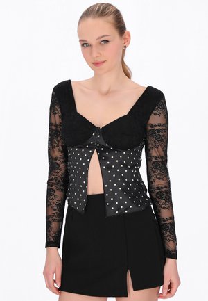Jonge vrouw die een zwarte top met witte stippen en kanten mouwen draagt, gecombineerd met een zwarte minirok met een frontale split, staand tegen een witte achtergrond.
