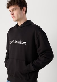 Juodas medvilninis džemperis su kengūrinėmis kišenėmis. Priekyje yra baltas "Calvin Klein" logotipas. Dizainą papildo pintas gobtuvas.
