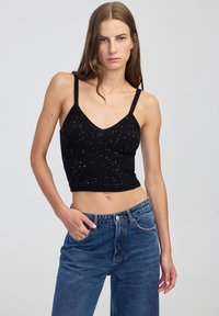 Žena v černém třpytivém crop topu a modrých džínech s vysokým pasem, stojící na pozadí jednoduchého světlého pozadí.