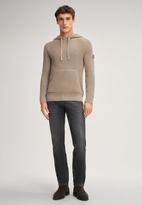 Haut en tricot beige avec une poche kangourou, des cordons de serrage, des poignets côtelés et un motif texturé, associé à un jean gris foncé et des bottes marron.