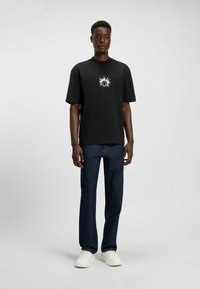 T-shirt en coton noir à manches courtes, arborant un graphique de soleil blanc sur la poitrine. Porté avec un jean foncé et des baskets blanches.