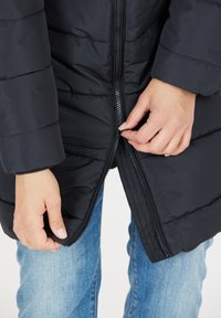 Mustapuffatakki, jossa on tikattu pinta ja vetoketju etupuolella, päällä vaaleansiniset farkut, kädet puristavat vetoketjua säätämistä varten.