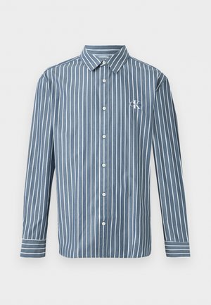 Camisa azul de manga larga con botones, con rayas blancas verticales y un pequeño logo blanco "CK" en el pecho izquierdo.