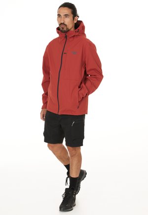 Rode waterdichte jas met een rits aan de voorkant, verstelbare capuchon en logo op de borst, gecombineerd met zwarte shorts en wandelschoenen.