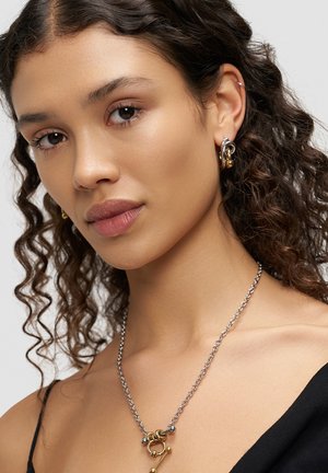 Lockiges Haar, trägt eine silberne Halskette mit goldenen Akzenten und einem Anhänger, kombiniert mit goldenen Creolen, vor einem neutralen Hintergrund.