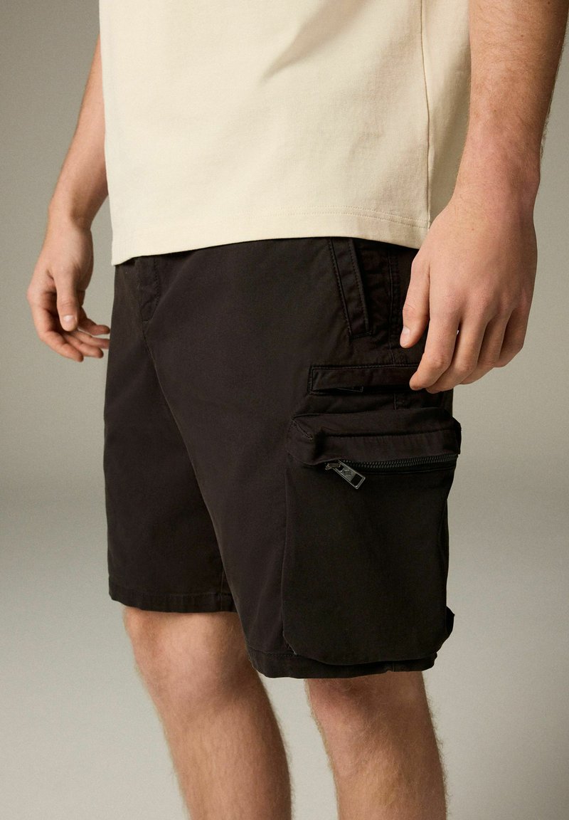 Shorts cargo noirs avec plusieurs poches, comprenant une poche zippée sur le côté, fabriqués en tissu léger, associés à un t-shirt beige.