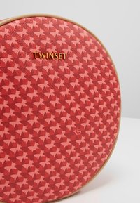 Okrągła torba z czerwoną teksturowaną powierzchnią, zdobiona różowym wzorem gwiazdek. Złote logo "TWINESET" i kremowe wykończenie.