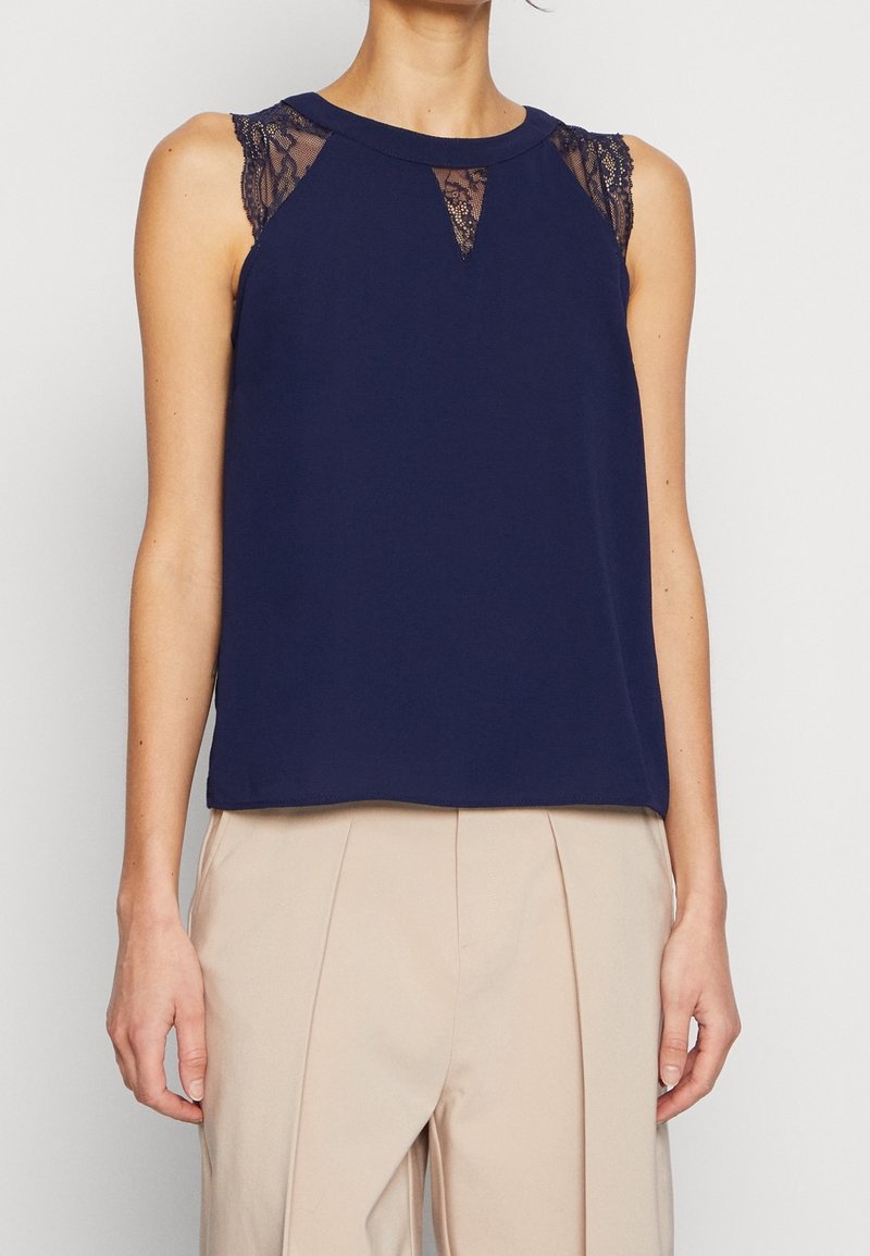 Haut sans manches bleu marine avec des détails en dentelle sur les épaules et le décolleté, associé à un pantalon plissé beige taille haute.