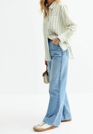 Vrouw draagt een lichtgroen gestreept overhemd met knopen, blauwe jeans met hoge taille en wijde pijpen, witte sneakers, houdt een kleine beige handtas vast.