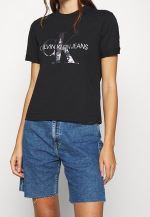 Osoba ubrana w czarny t-shirt Calvin Klein Jeans z logo oraz niebieskie dżinsowe szorty z surowym wykończeniem, stojąca na białym tle.