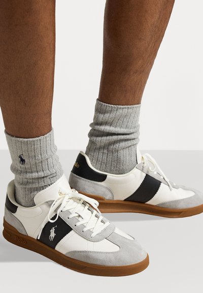 Adidași albi și gri cu dungi negre, detalii din suede texturat și tălpi din cauciuc maro, asortați cu șosete gri cu model ribetat, având un logo.