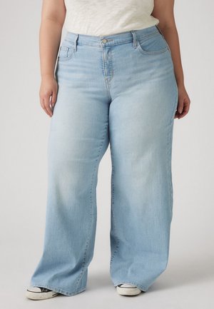 Personne portant un jean bleu clair à jambes larges et des baskets blanches, debout devant un fond uni.