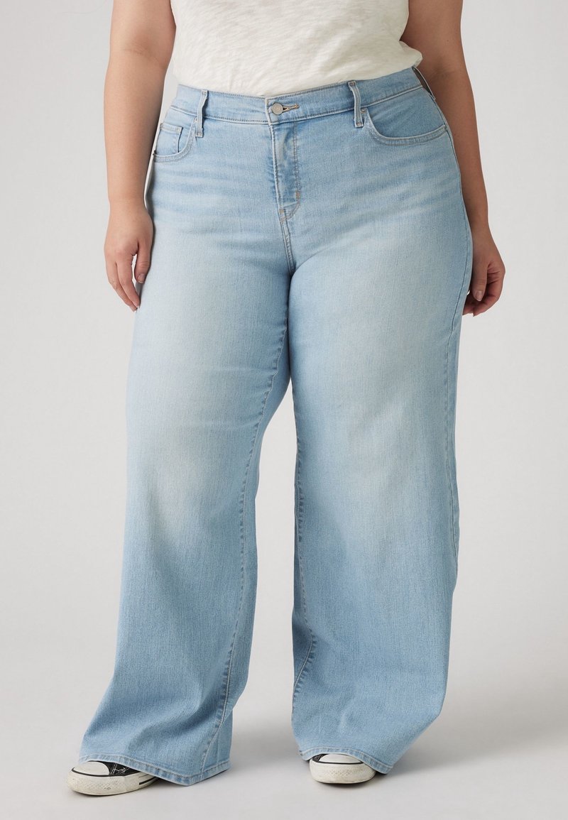 Personne portant un jean bleu clair à jambes larges et des baskets blanches, debout devant un fond uni.