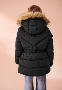 Černá prošívaná bunda s kapucí lemovanou umělým kožíškem, páskovaným pasem a chevronovým prošíváním. Látka vypadá hladká a izolovaná.