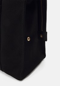 Coin d'un sac en tissu noir avec rivet métallique doré visible et fermeture éclair noire partielle avec dents dorées sur fond blanc.