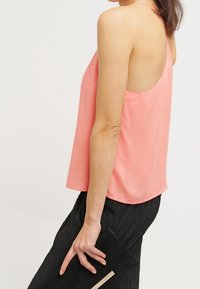 Mujer con una blusa sin mangas color coral y falda negra sosteniendo un bolso de mano negro frente a un fondo blanco liso.