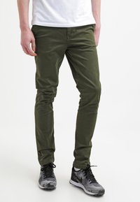 Olivgröna slim-fitbyxor i bomullstwill. Slät textur, femficksdesign, tillsammans med grå och svarta träningsskor.