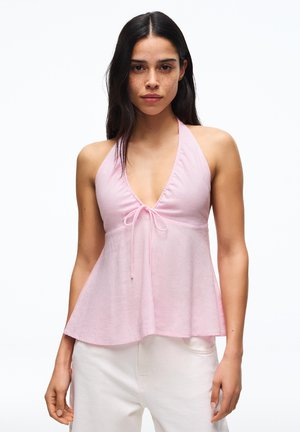 Hosszú sötét hajú nő világos rózsaszín ujjatlan halter felsőt visel elöl megkötve és fehér nadrágot, előre néz fehér háttér előtt.