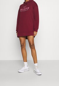 Vestido sudadera burdeos con logo frontal, capucha con cordón y puños de canalé, combinada con zapatillas altas blancas y calcetines tobilleros.