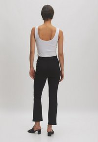 Weißes ärmelloses Top, schwarze, figurbetonte Hose mit ausgestellt Fuß. Das Modell trägt schwarze Absatzmules; glattes Material ohne sichtbare Muster.