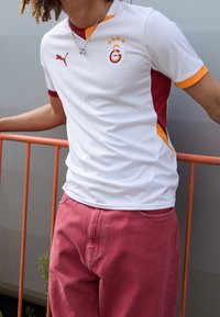 Vit fotbollströja med röda och orangea detaljer, geometriskt mönster, broderad logotyp och tre guldstjärnor ovanför. Korta ärmar.