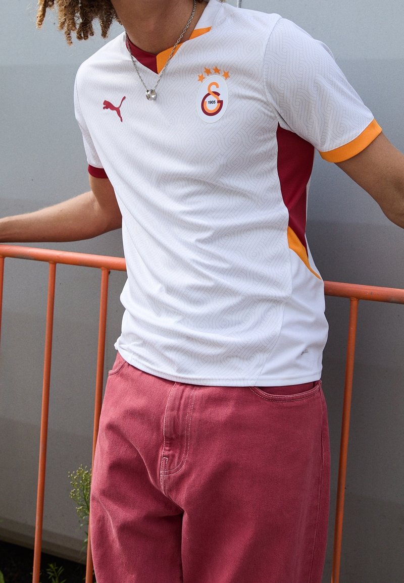 Vit fotbollströja med röda och orangea detaljer, geometriskt mönster, broderad logotyp och tre guldstjärnor ovanför. Korta ärmar.