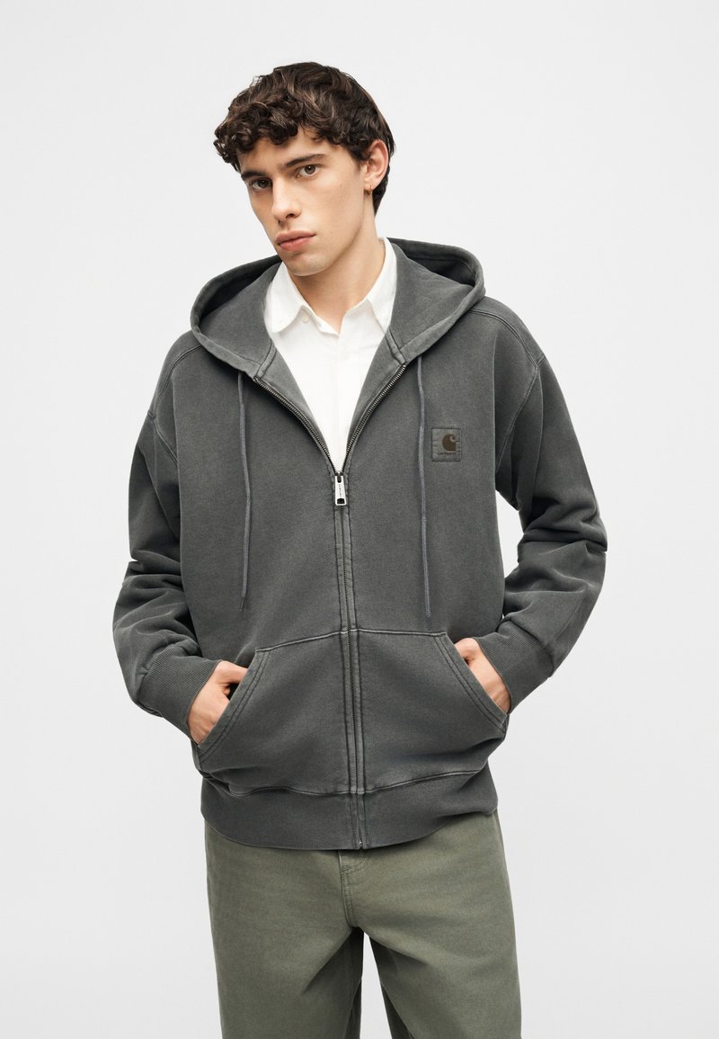 Young man wearing a dark gray zip-up hoodie over a white button-up shirt with hands in front pockets, standing against a plain background.