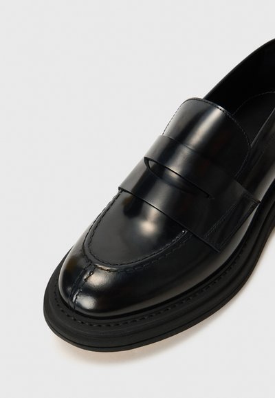 Mocassins en cuir noir avec une finition brillante, dotés d'une large sangle brillante sur le dessus et d'une semelle texturée pour l'adhérence.
