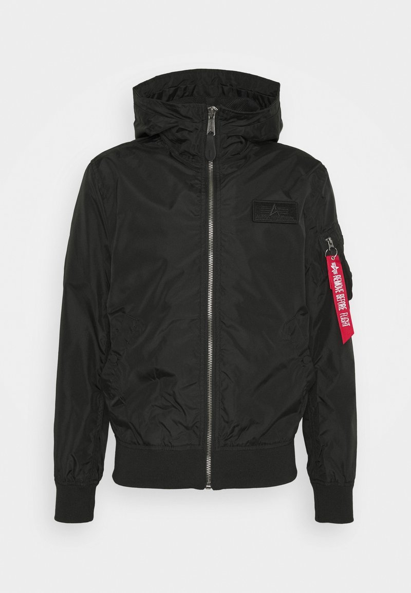 Veste bomber noire zippée à capuche avec poignets côtelés, patch sur la poitrine et étiquette rouge "Remove Before Flight" sur la fermeture éclair de la poche de la manche gauche.