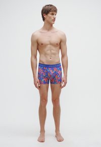 Eng anliegende Herren-Badehosen mit blauem Grund und buntem Blumenmuster. Weicher Stoff mit einem Taillenbund mit Markenlogo.