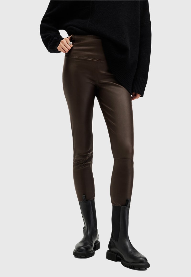 Osoba ubrana w ciemno-brązowe, skórzane legginsy z wysokim stanem, czarne masywne botki i luźny czarny sweter, z jedną ręką na biodrze.