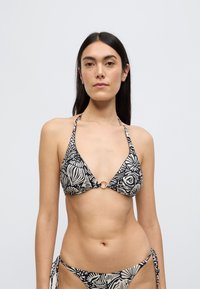 Σετ μπικίνι σε μαύρο και εκρού με τοπ με σχέδιο, χιαστί στο λαιμό, με τριγωνικά κοχύλια και χρυσό δαχτυλίδι στο κέντρο, και κάτω μέρος με αντίθεση στο σχέδιο.