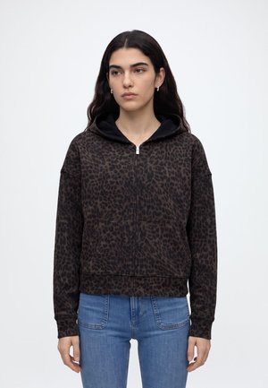 Junge Frau mit langen dunklen Haaren, die einen dunkelbraunen Kapuzenpullover mit Leopardenmuster und blaue Jeans trägt und leicht zur Seite schaut.