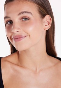 Model met lang bruin haar, gekleed in een zwarte top. Heeft gouden studs in de oren. De huid heeft een gladde, stralende textuur; de lippen zijn een zachte nude tint.
